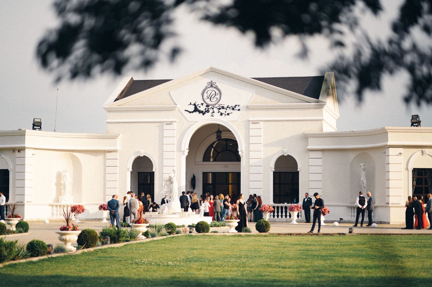 Elegant white neoclassical building entrance with columns and archways surrounded by manicured lawn and visitors.