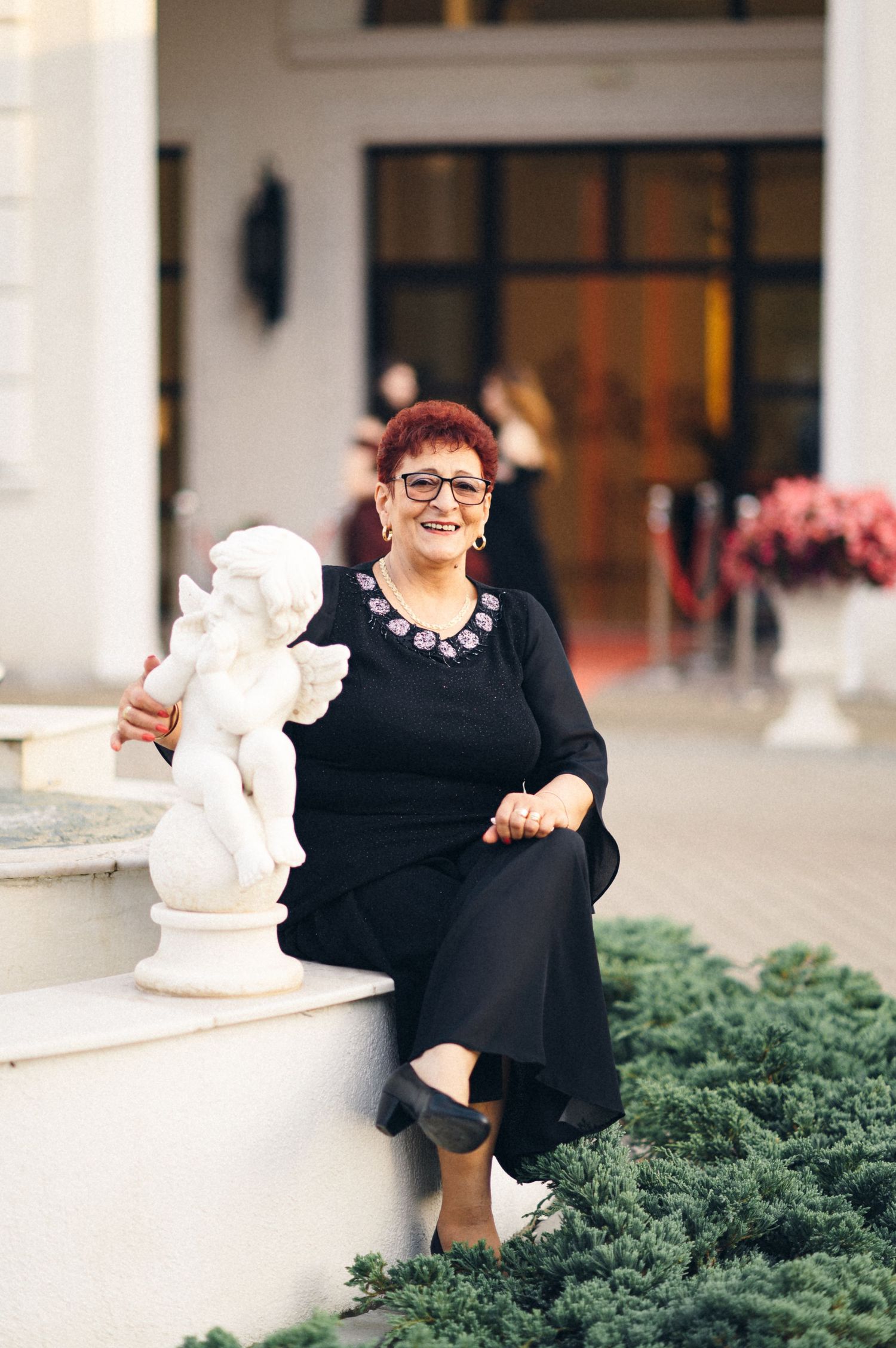 A person in black attire sits beside a white decorative statue outside an elegant building entrance.