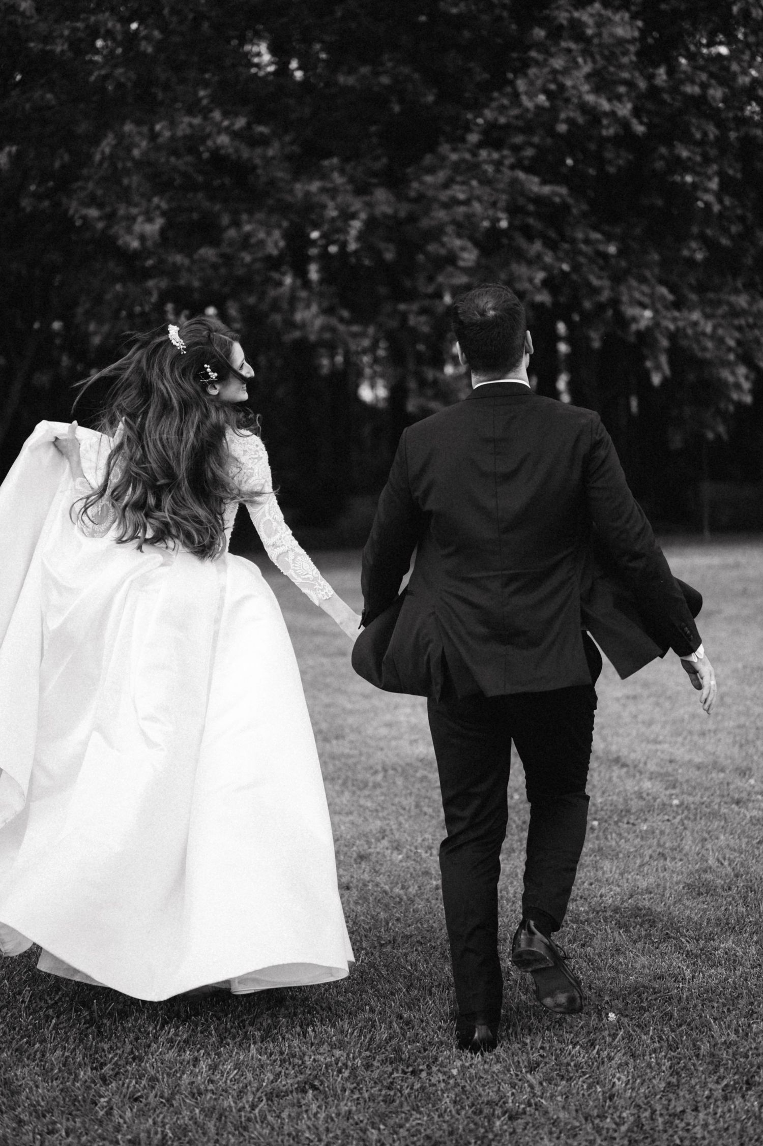A couple in wedding attire runs playfully across a grassy lawn in black and white photography.
