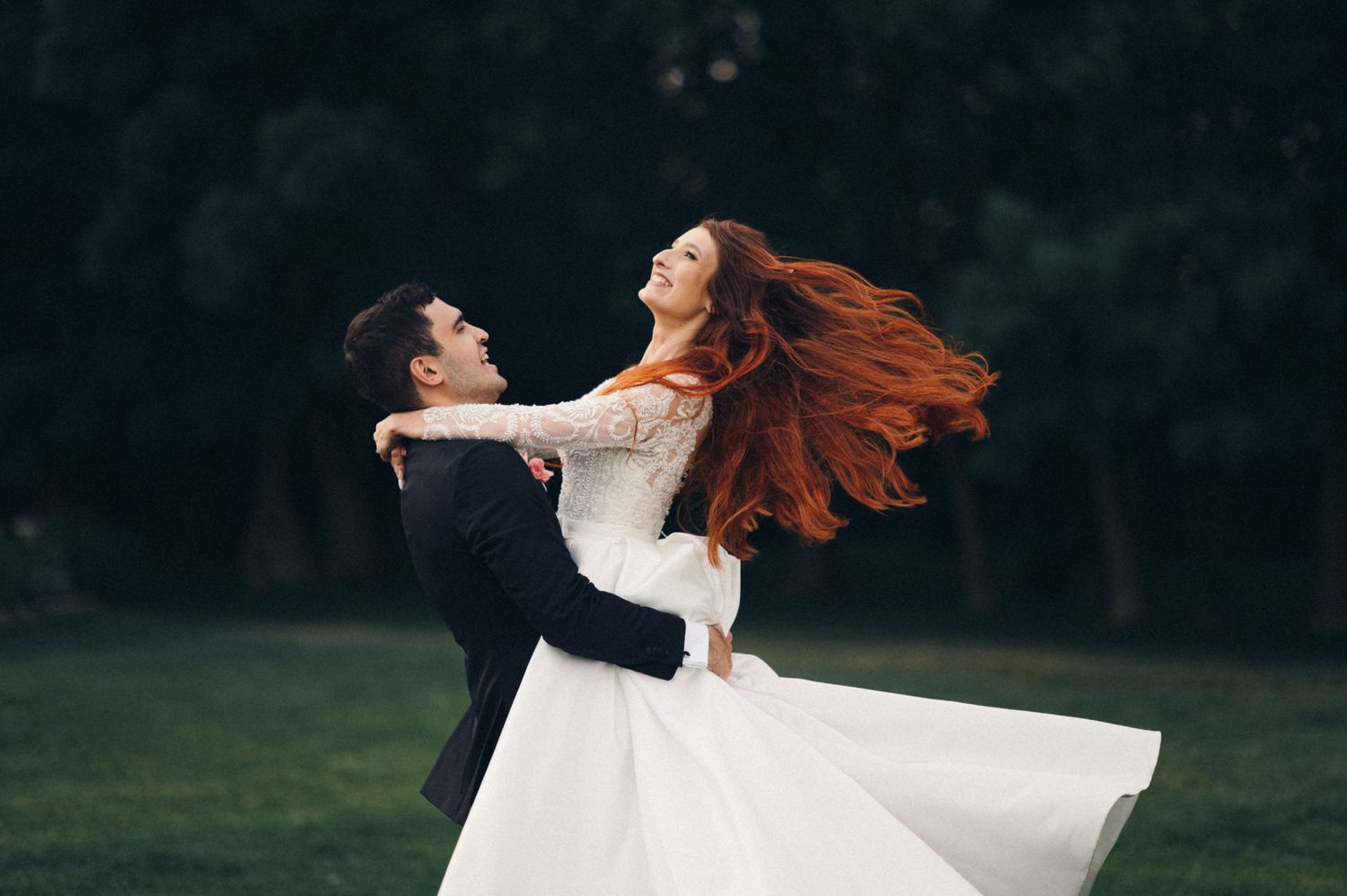 A couple in wedding attire dances on grass, with the bride's red hair and white dress flowing dramatically.