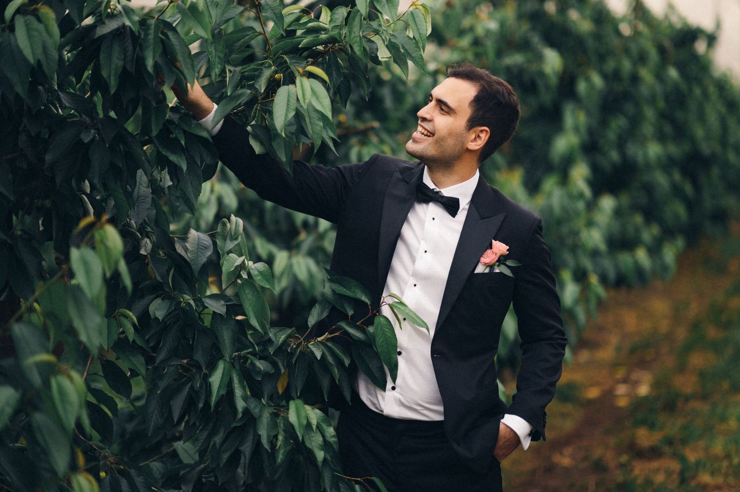 A groom in classic black tuxedo poses elegantly among garden foliage.