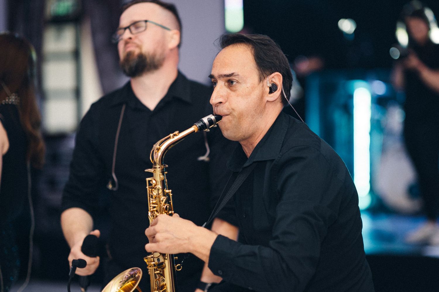 Saxophone player in black attire performs with band members at an indoor event.