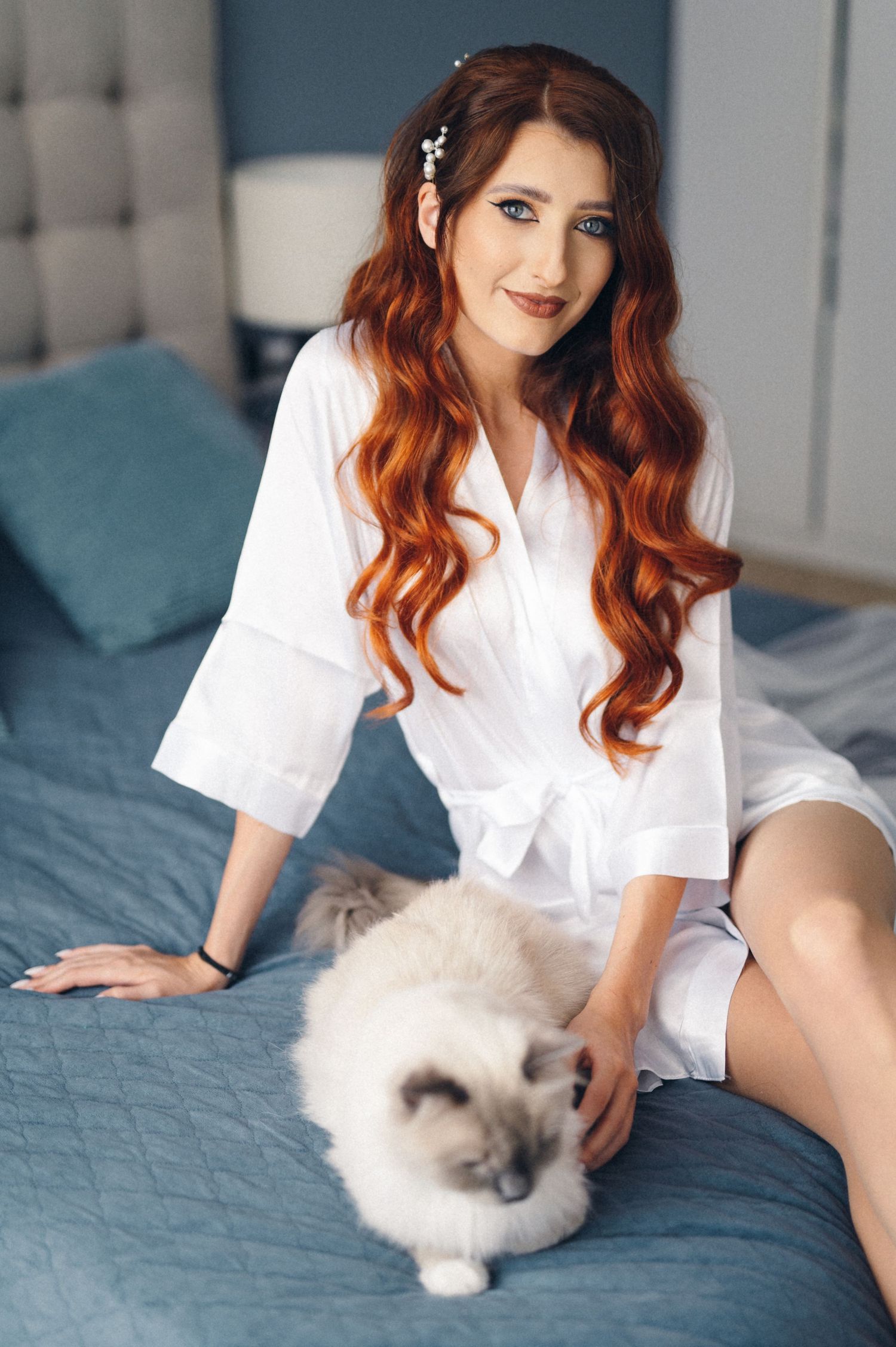 Long wavy red-haired figure in white robe sitting on blue bed with fluffy white cat.