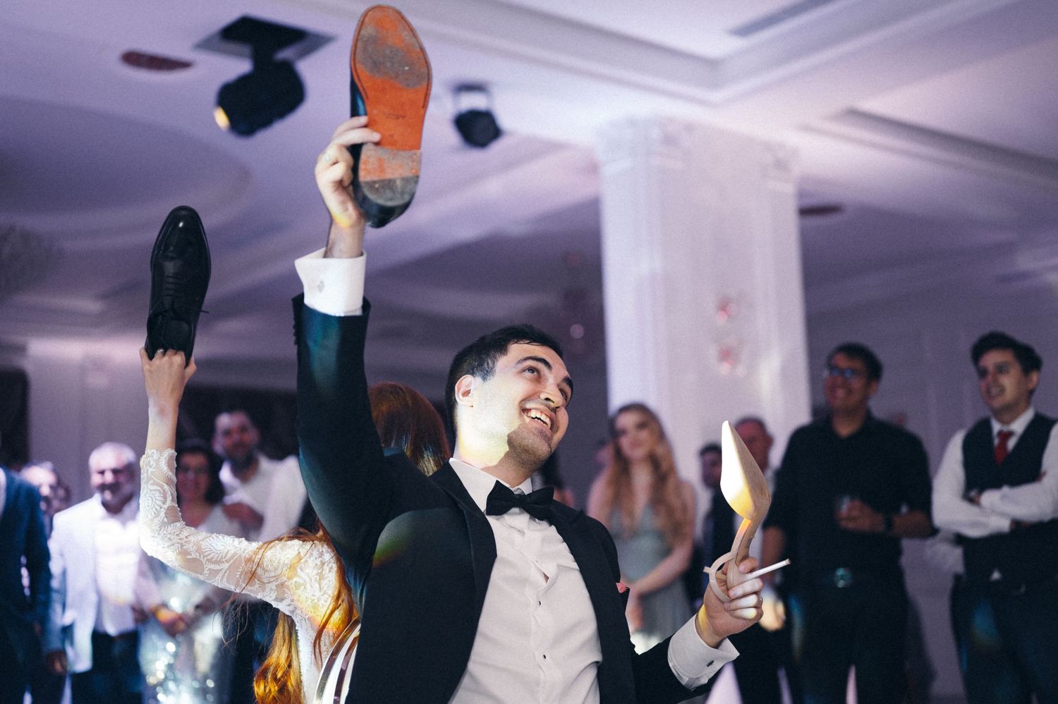 Wedding guests hold up their shoes during a lively dance moment at an indoor reception celebration.