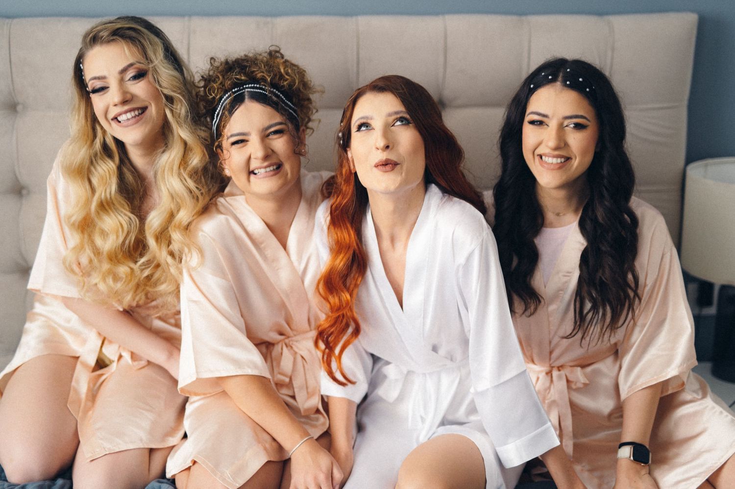 Four bridesmaids in silk robes sitting together on bed sharing laughter.