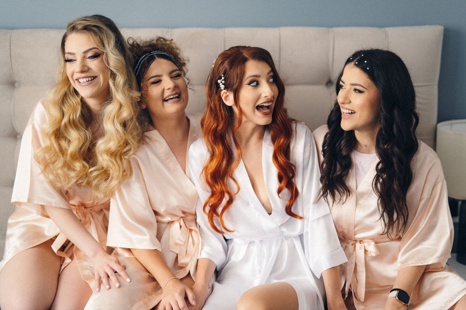 Candid moment of four bridesmaids in matching robes sharing jokes on bed.