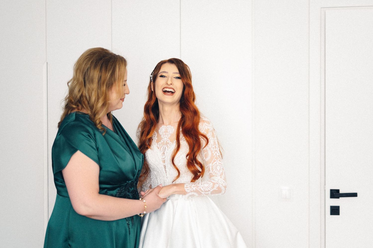 A bride in white lace dress shares a joyful moment with someone in a teal dress.