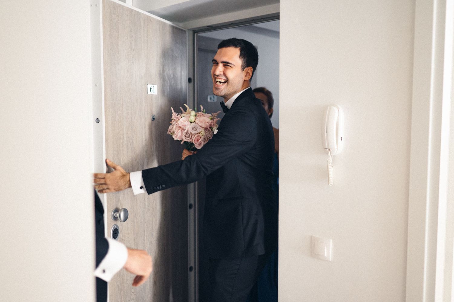 Person in dark hoodie smiling while holding something at a doorway.