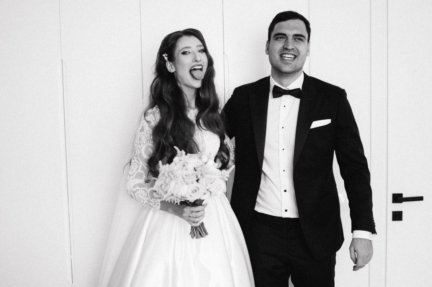 Black and white portrait of newlyweds laughing together against white wall.