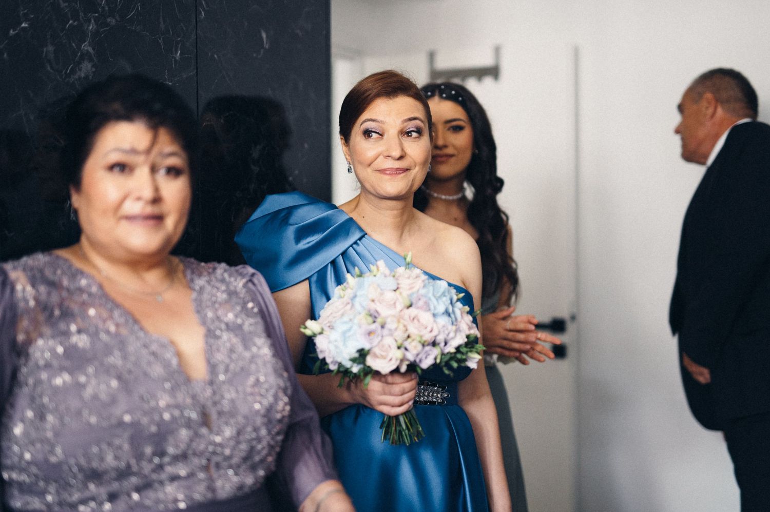 Wedding party members in formal attire holding bouquets in preparation room.