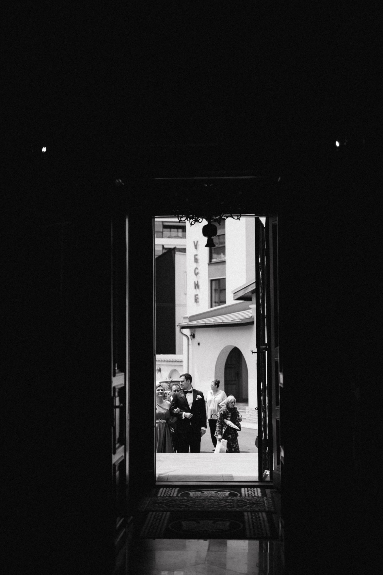 Black and white view through doorway of formal wedding procession outside building.