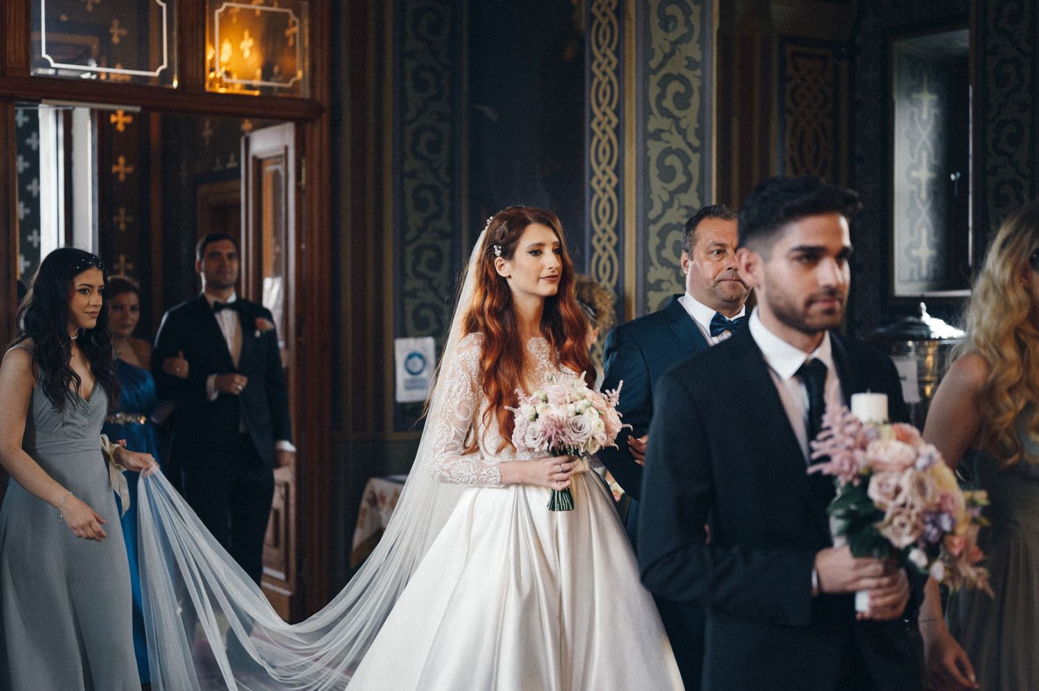 Elegant wedding party stands in a beautifully decorated church with ornate walls and lighting.