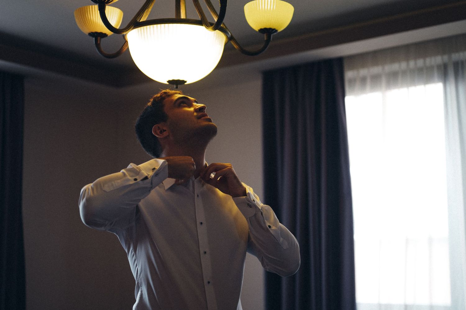 A person adjusts their tie while looking up in a dimly lit room with a vintage chandelier.