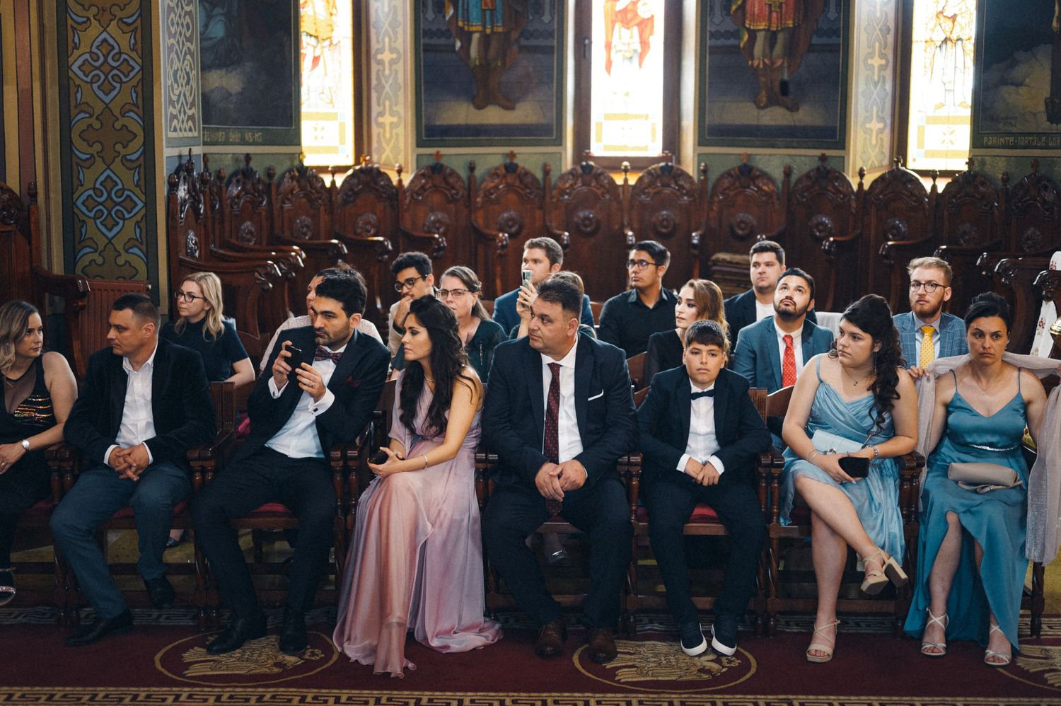 Wedding guests seated in formal attire inside historic church with painted walls.