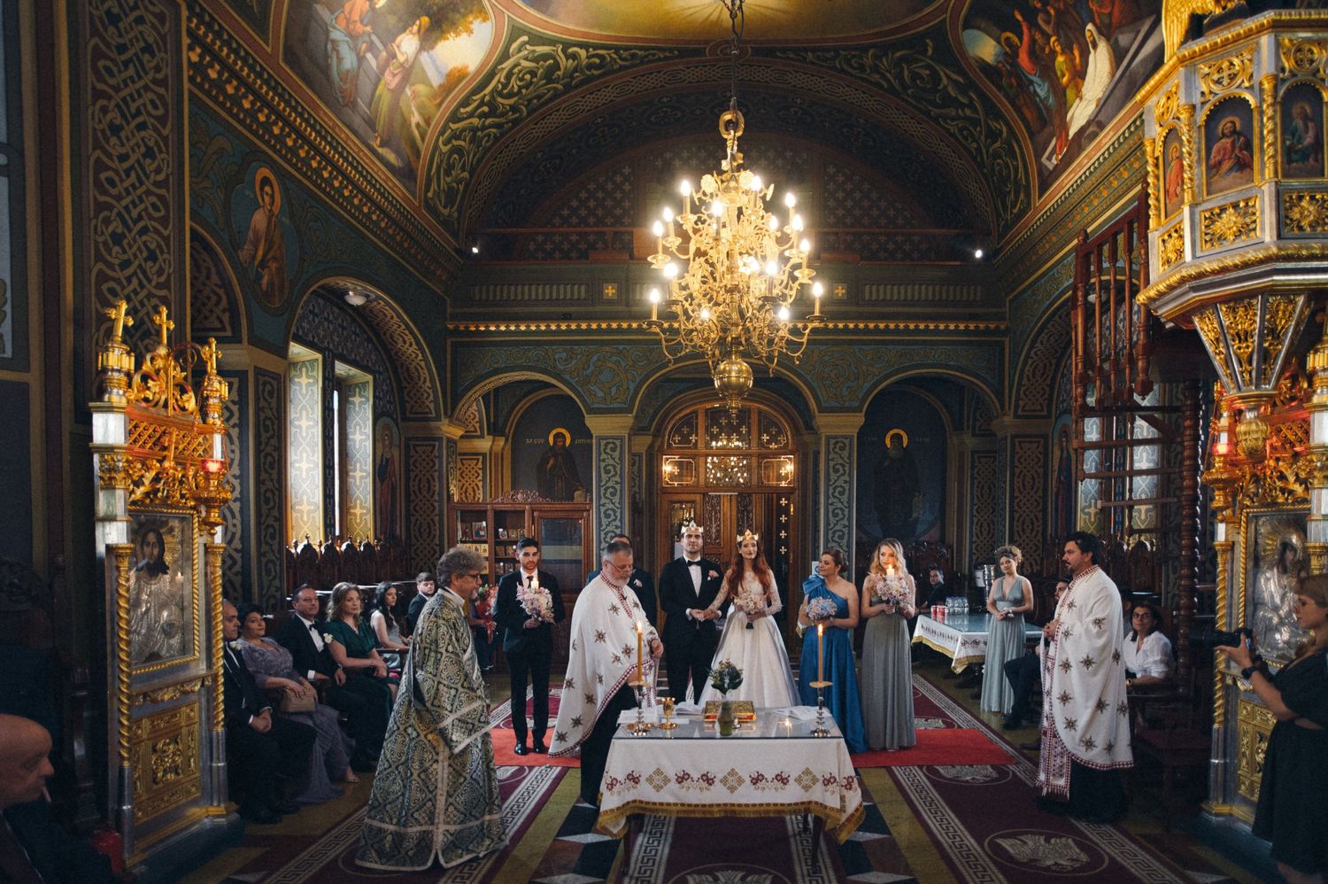 Interior of grand Orthodox church during wedding ceremony with golden chandeliers and decorated altar.