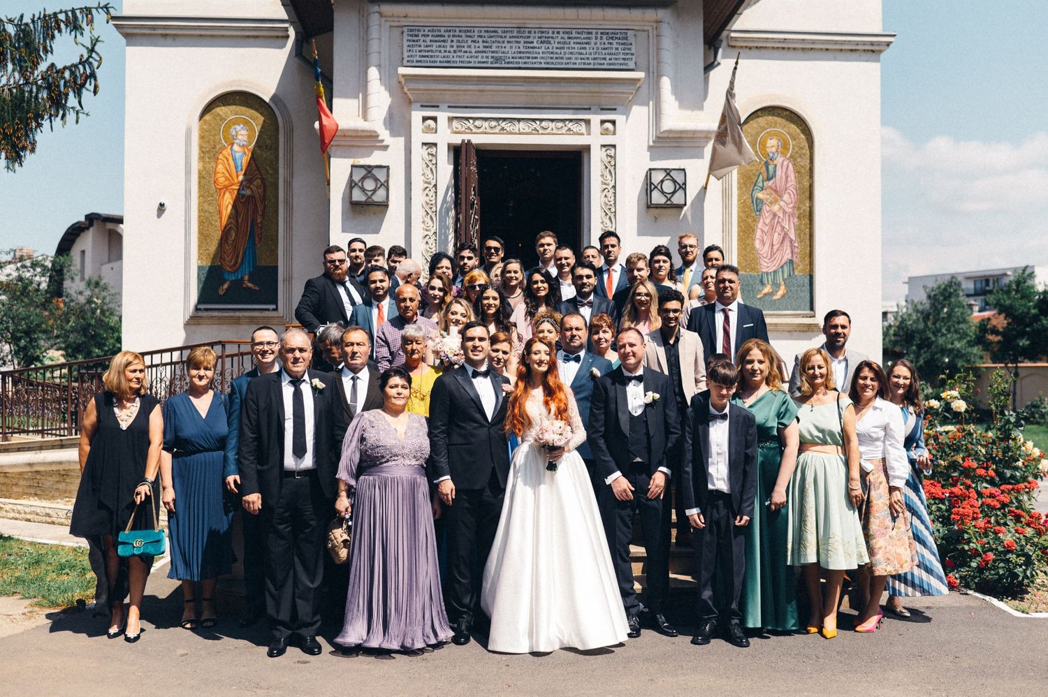Large group photo of wedding party and guests gathered outside Orthodox church entrance.