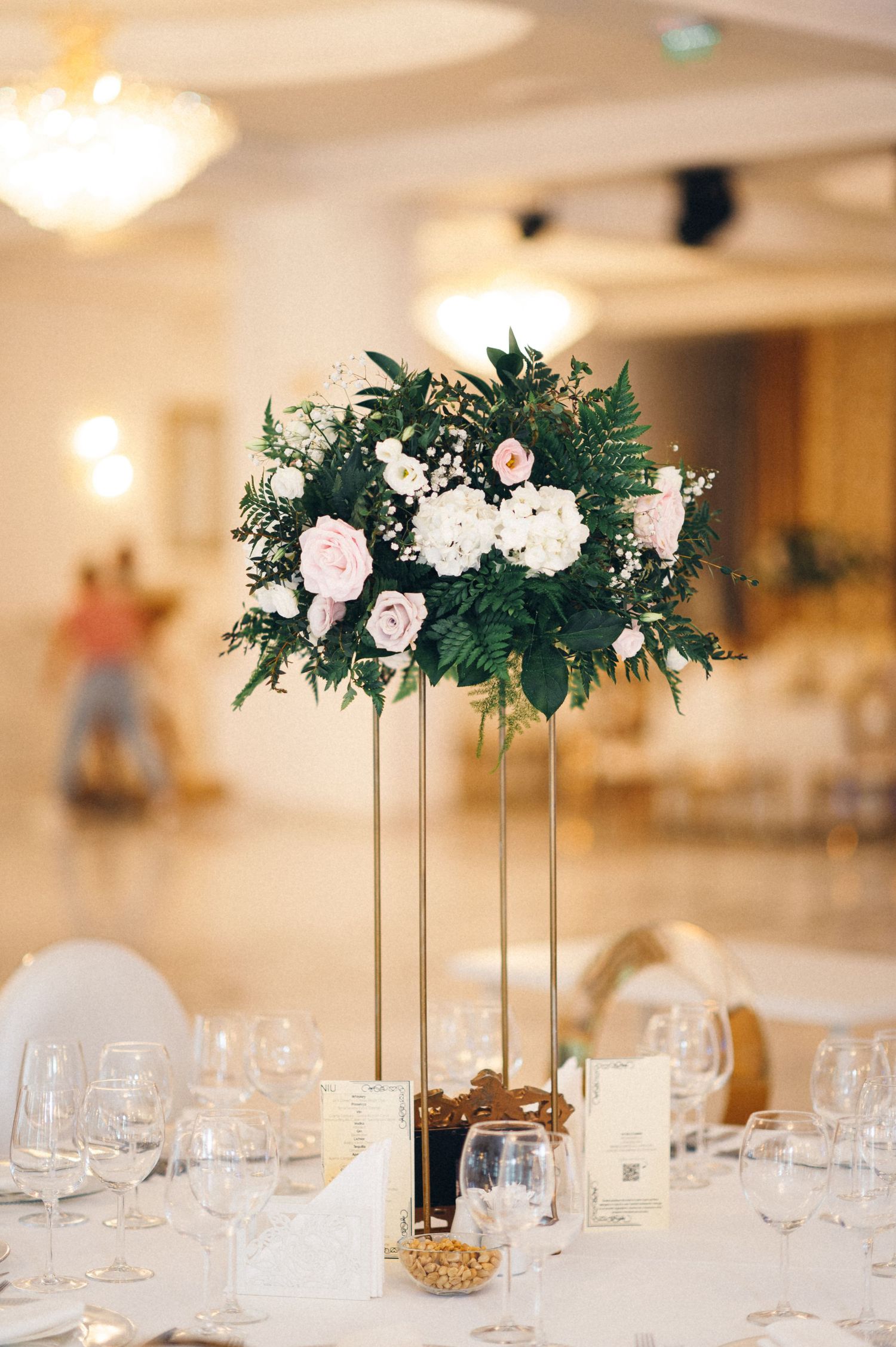 Tall floral centerpiece with white and pink roses mixed with ferns on a gold stand at an elegant reception table.