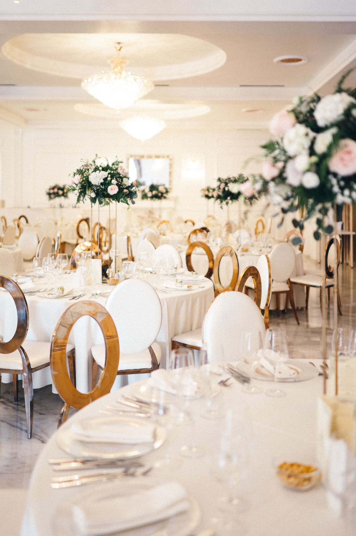 Luxurious wedding reception hall with white tables, gold chairs, and crystal chandeliers.