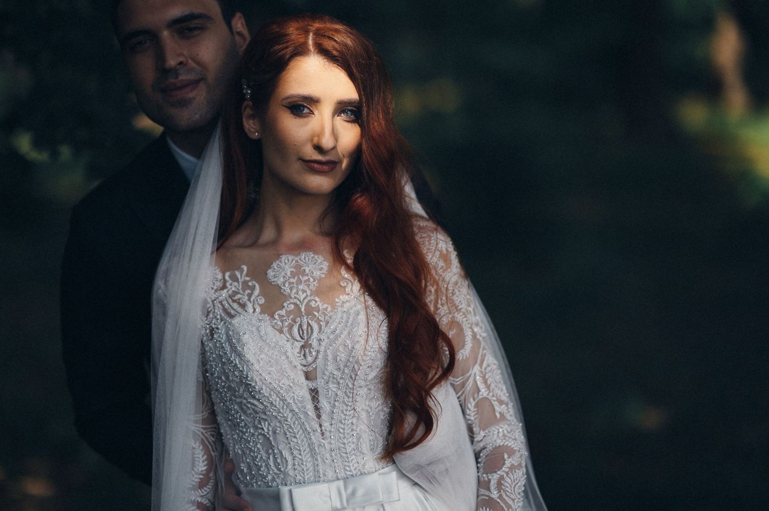 An elegant bridal portrait highlighting intricate lace details and flowing red hair against natural foliage.