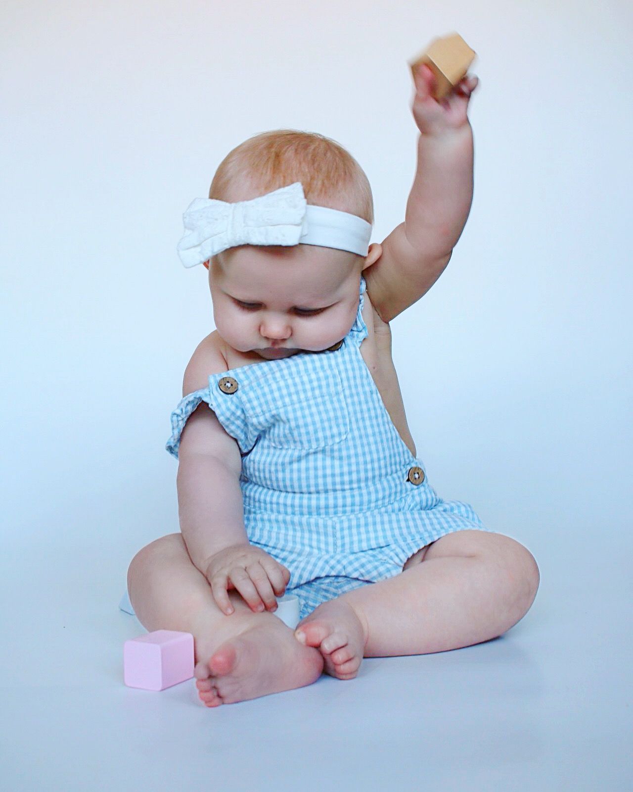 Baby in blue gingham romper and white headband playing with toys on white background.