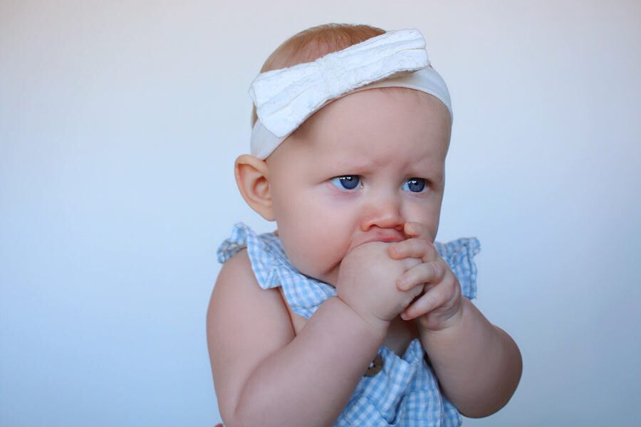 Baby wearing white headband and blue outfit putting fingers in mouth.