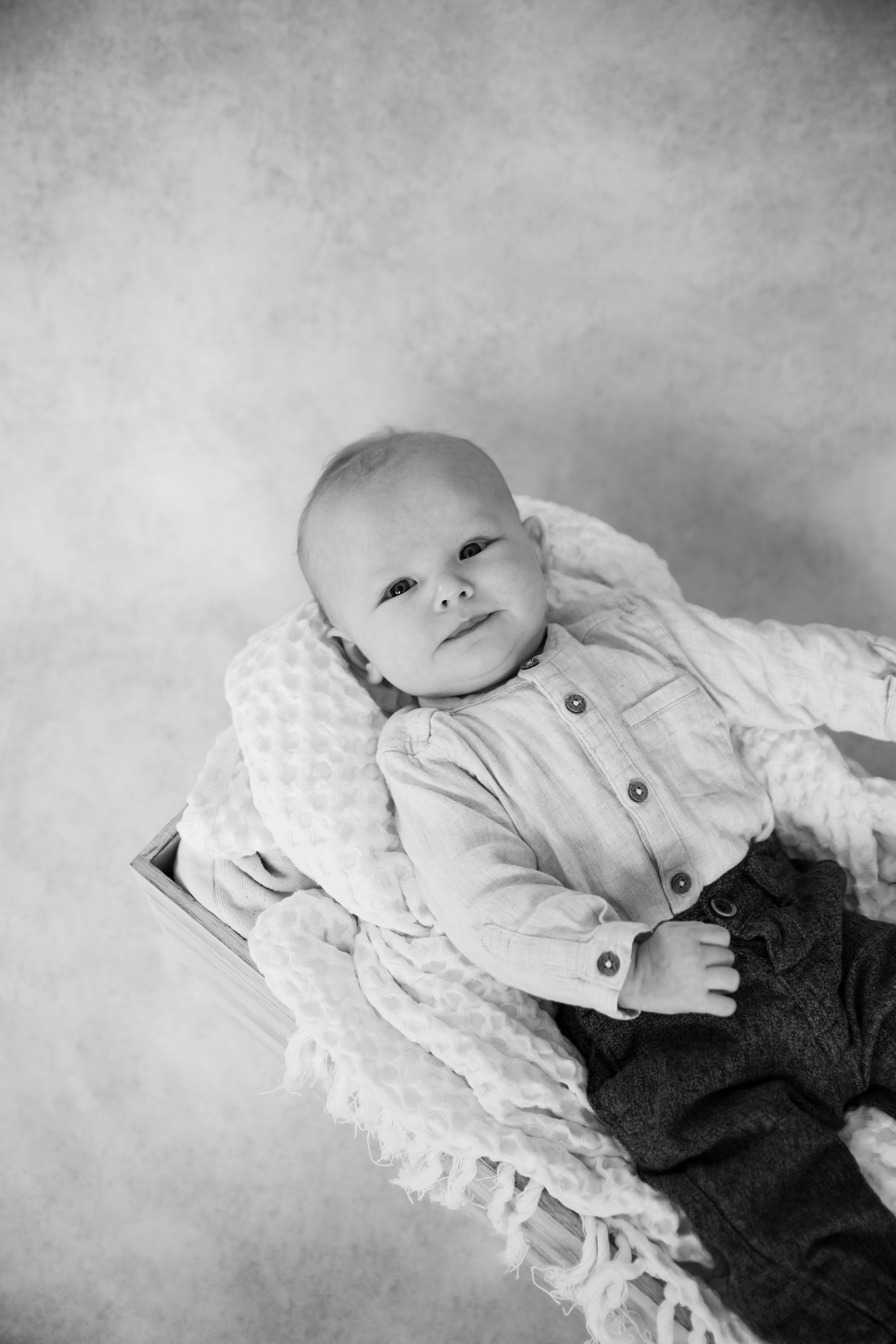 Black and white portrait of a baby in buttoned clothing on lace-trimmed fabric.