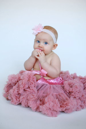 Baby wearing pink tutu and white headband sitting against white backdrop.