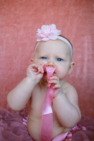 Baby in pink attire with flower headband playing with a pink ribbon against coral backdrop.