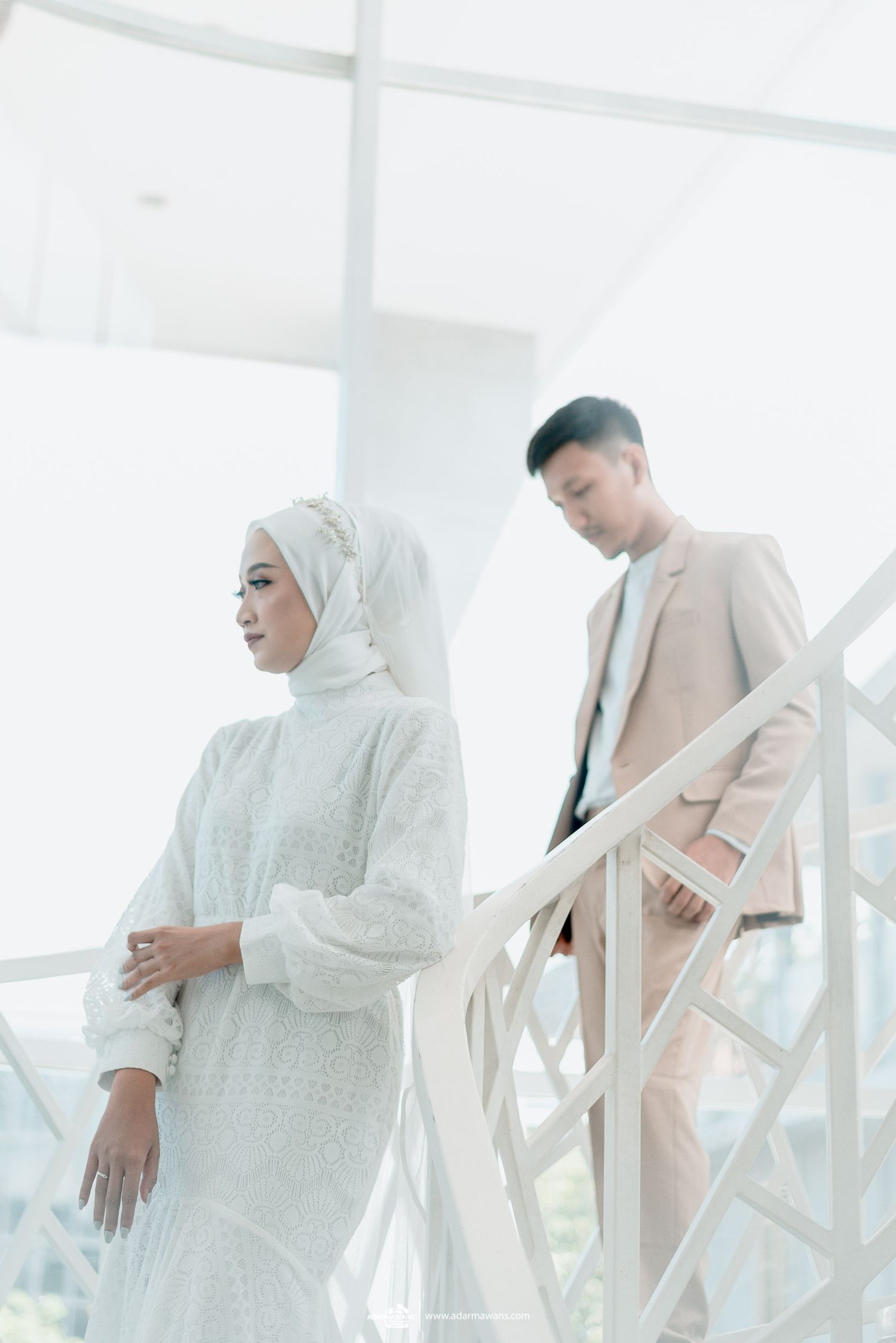 Elegant couple in beige and white attire pose by bright window in modern setting.