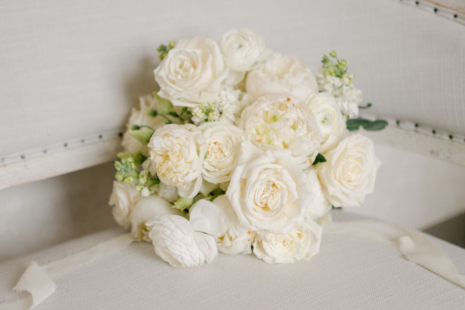 Lush white rose and ranunculus bridal bouquet resting on a white tufted chair for a classic wedding.