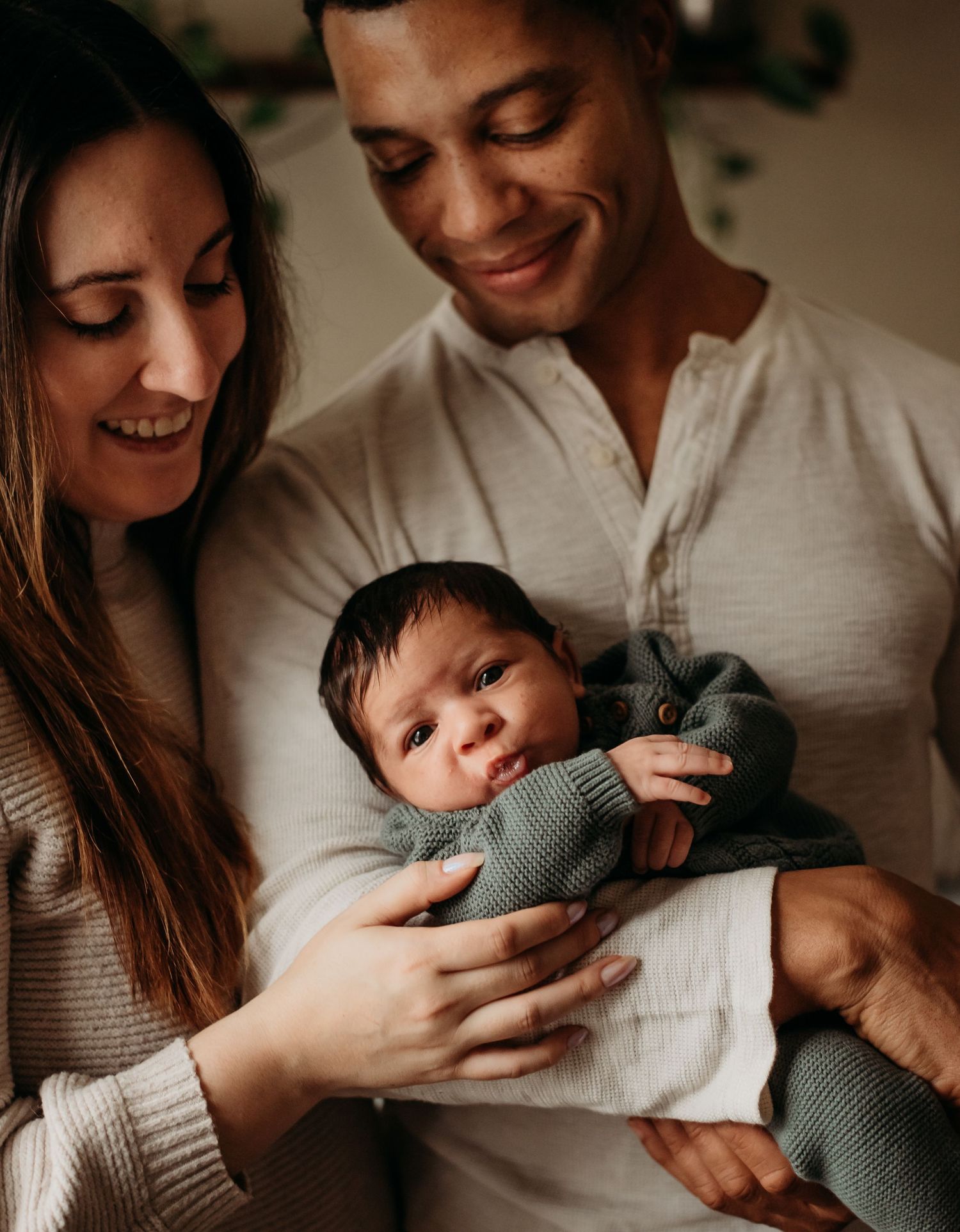 Smiling parents tenderly hold their newborn baby dressed in a sage green knit outfit.