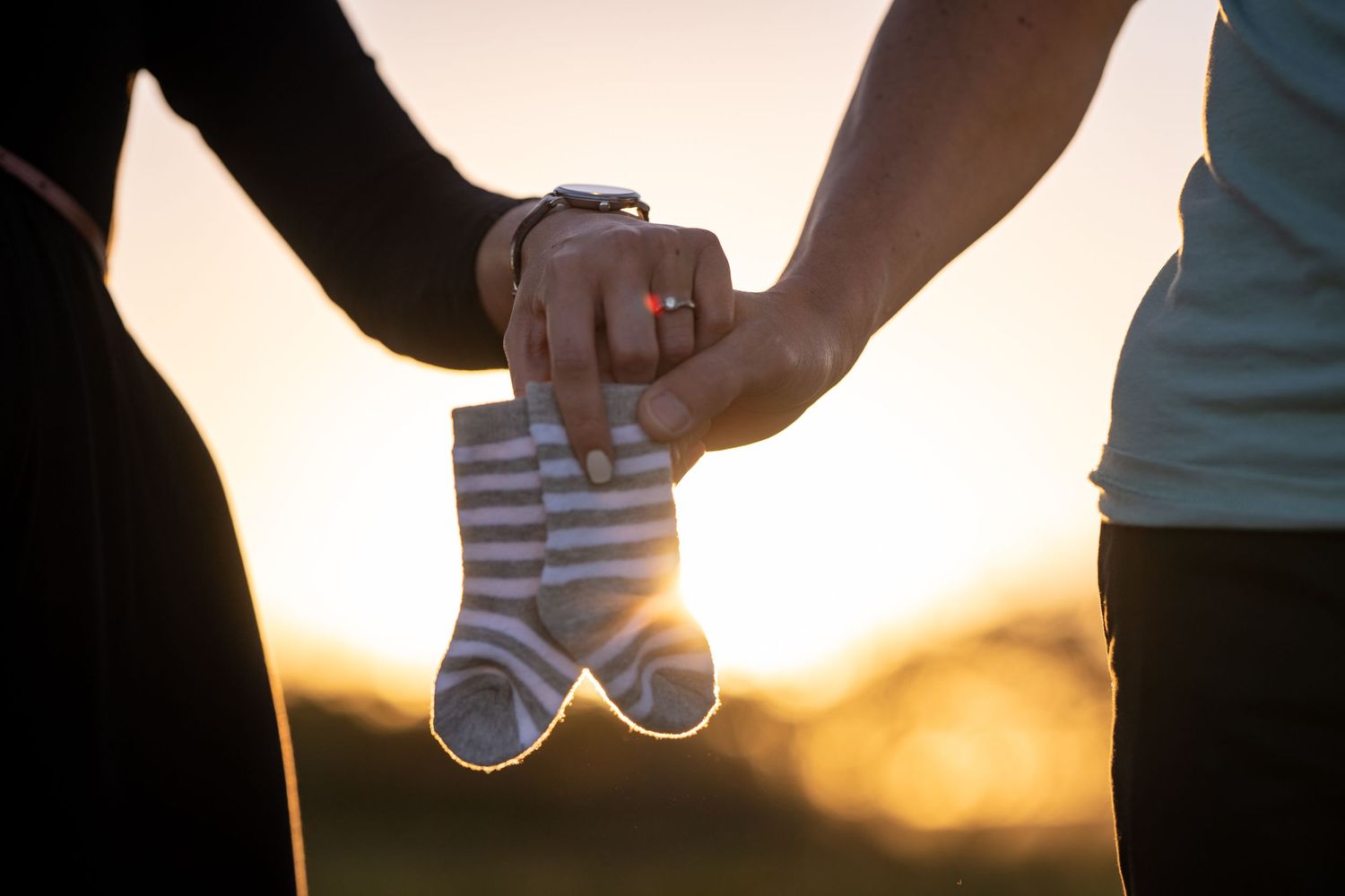 Couple holding tiny baby socks together at sunset, announcing pregnancy.