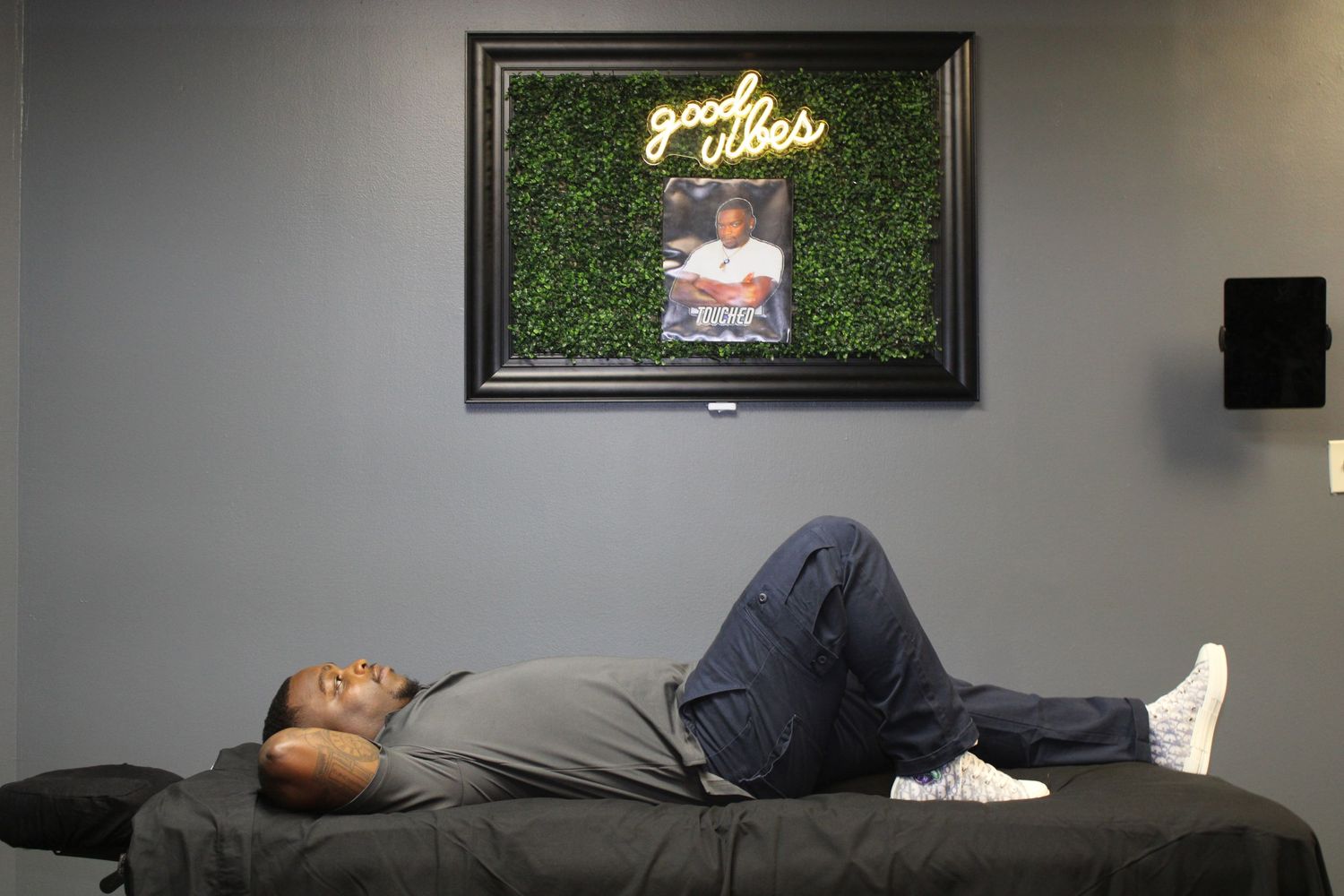 Person relaxing on a black couch beneath a neon sign and greenery wall display.