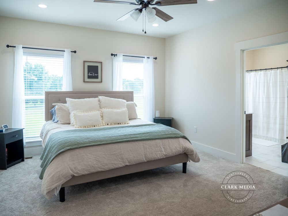 Bright master bedroom with gray bedding, ceiling fan, and large windows.