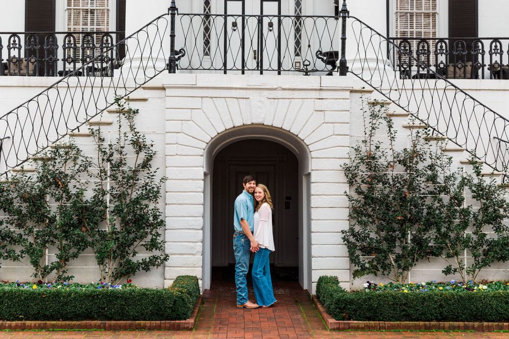A couple standing in an elegant white archway entrance with symmetrical landscaping.