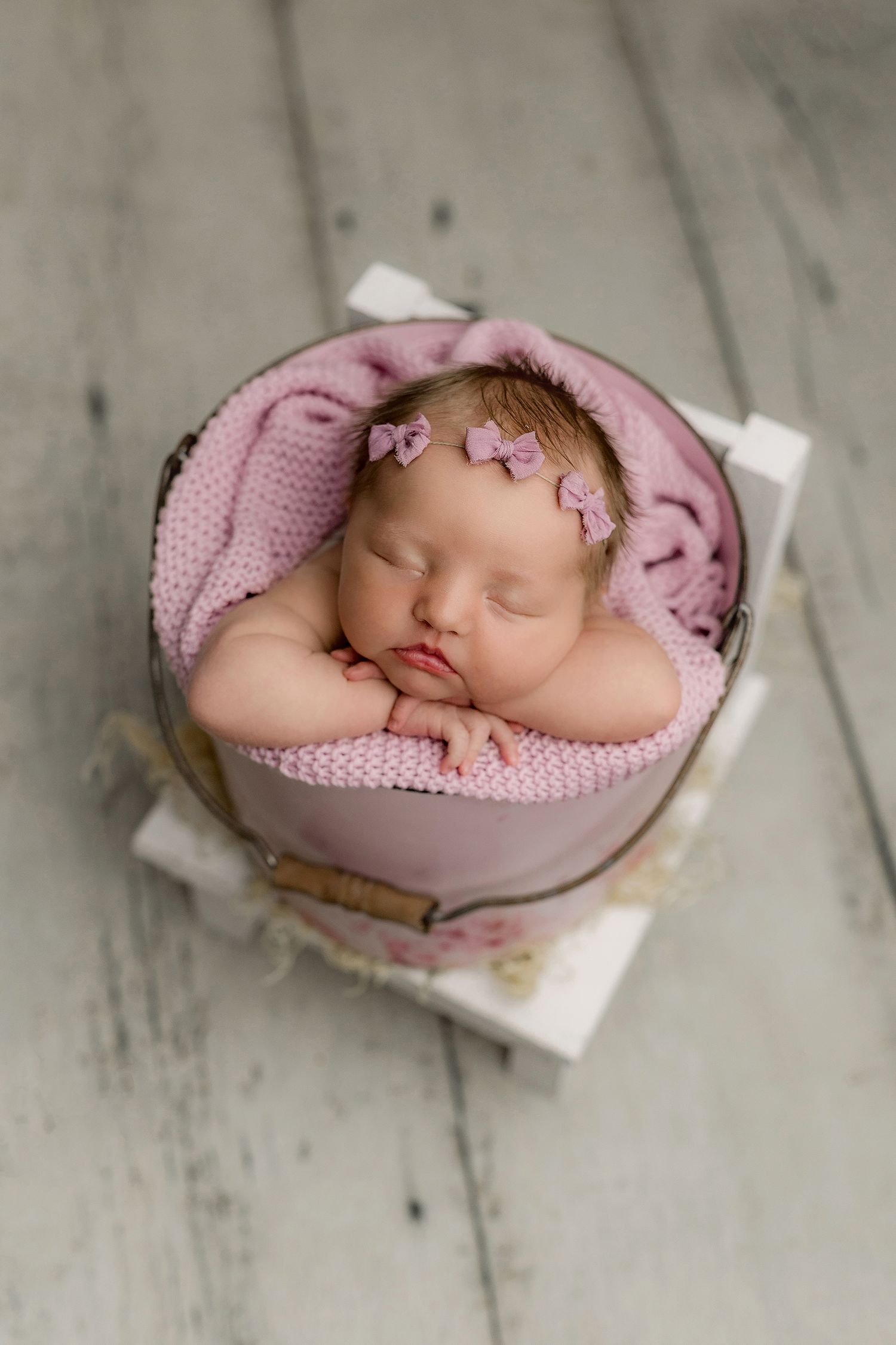 newborn baby girl in pink bucket on little chair with head on hands with pink bow