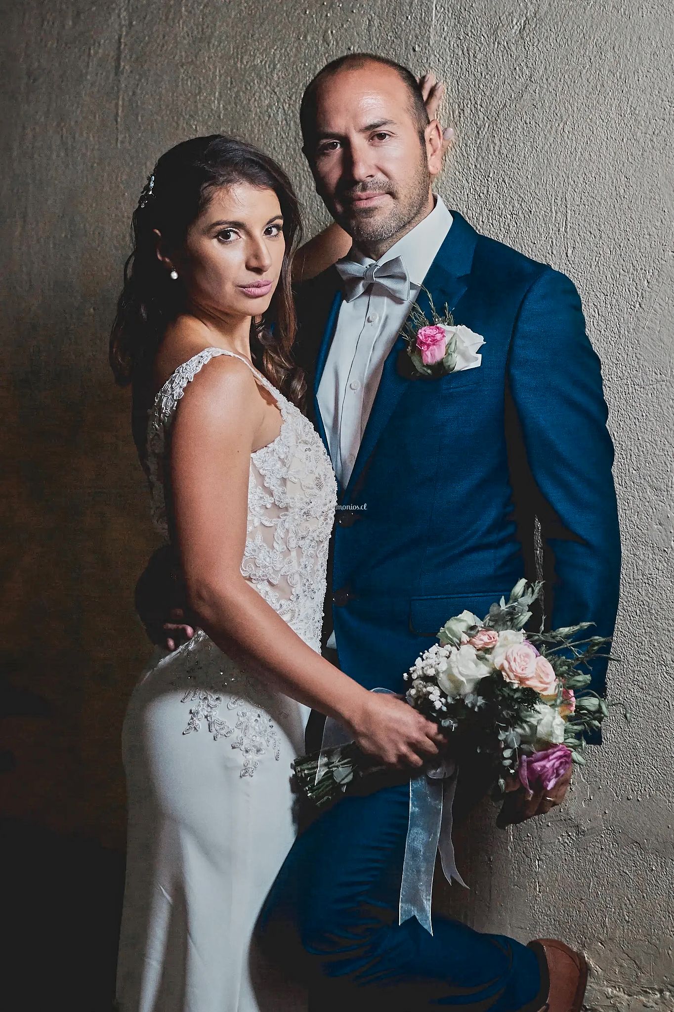 Elegant wedding couple poses against textured wall, bride in lace gown and groom in navy suit.