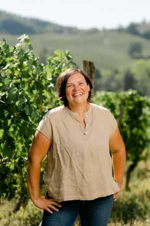 Person standing in sunny vineyard with grapevines and rolling hills in background.