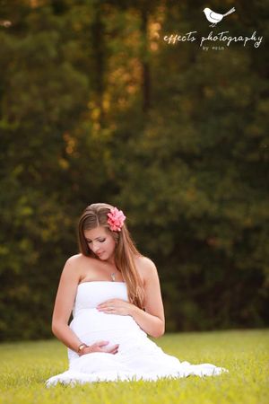 Ethereal maternity portrait of woman in white dress sitting on grass with sunlit forest background.