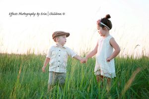 Two children in vintage-style clothing stand together in a tall grassy field at sunset.