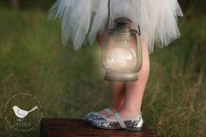A lantern glows softly next to small feet in ballet flats standing on a wooden surface with tulle skirt visible.