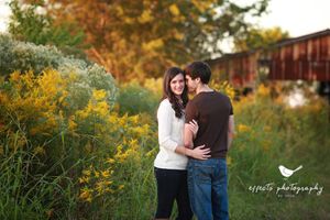 Romantic couple embracing among yellow wildflowers with rustic train car in background at sunset.