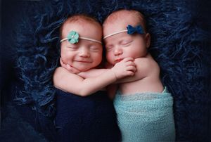 Twin newborns wearing headbands cuddling together on blue textured backdrop.