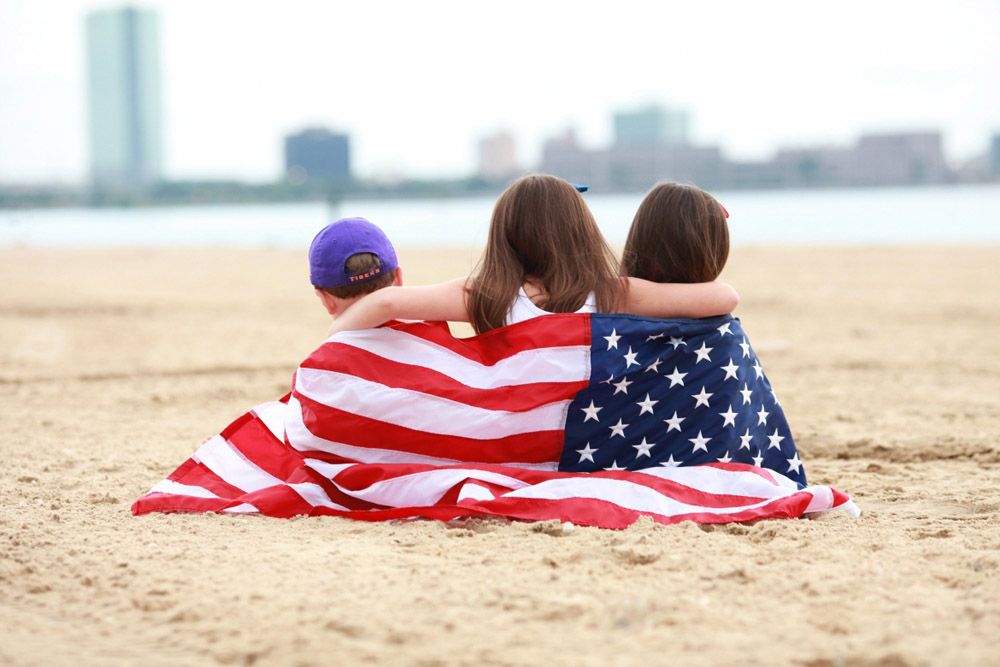 Three children wrapped in American flag sitting on beach looking at skyline.