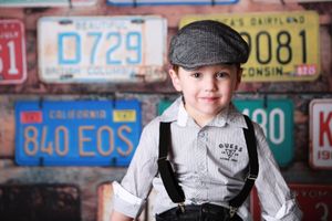 Vintage-styled photo against wall of old license plates featuring child in suspenders and newsboy cap.