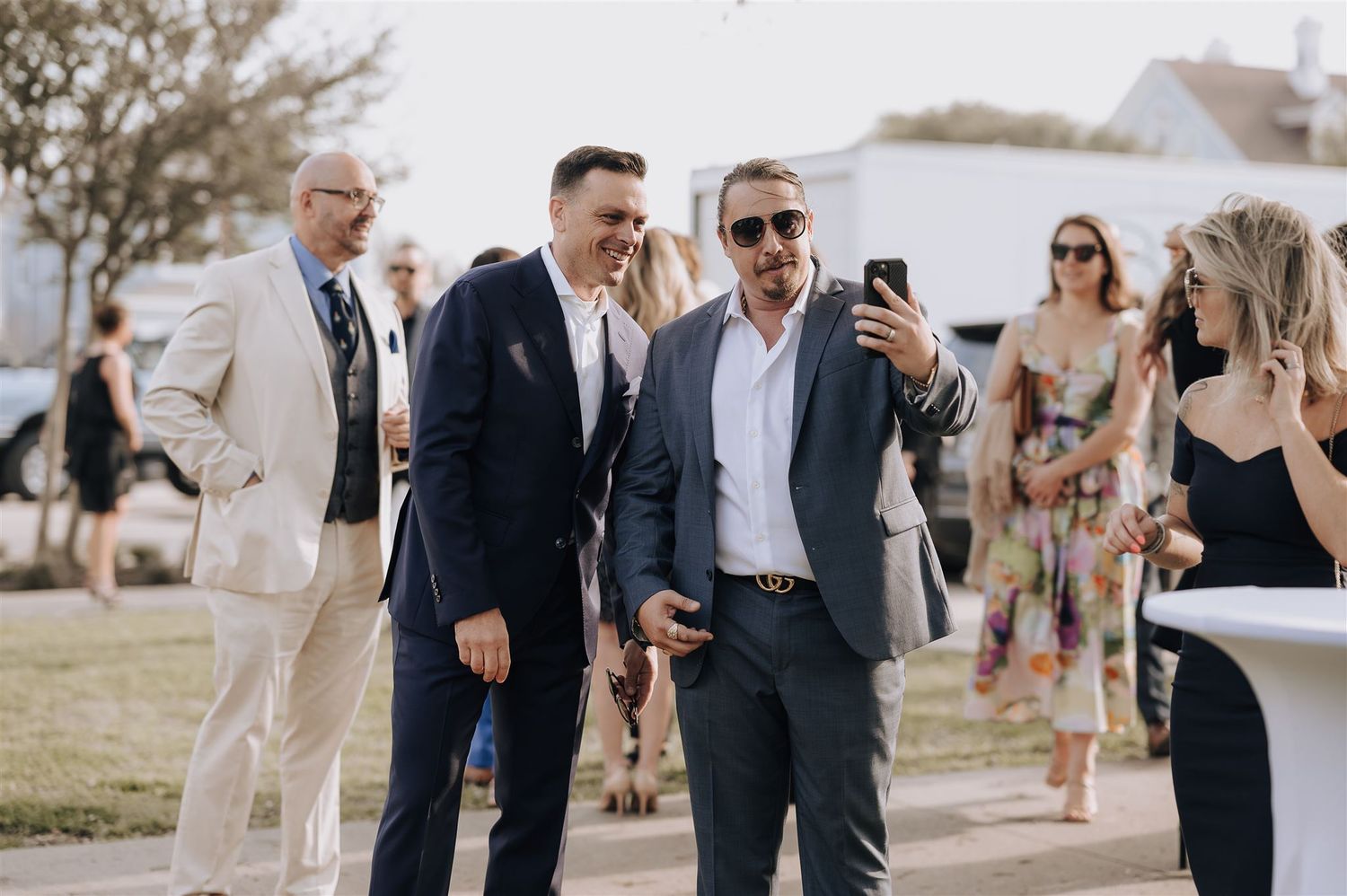 Guests in formal attire share a moment while taking selfies at outdoor wedding venue.