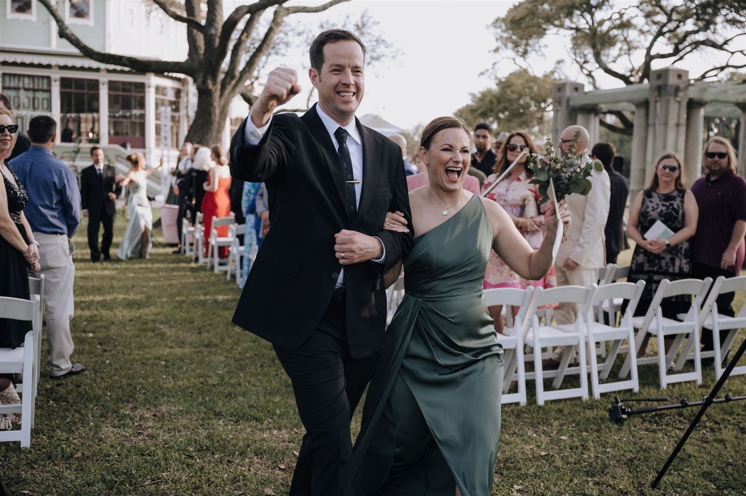 Wedding attendees celebrate with joyful gestures while walking across lawn.