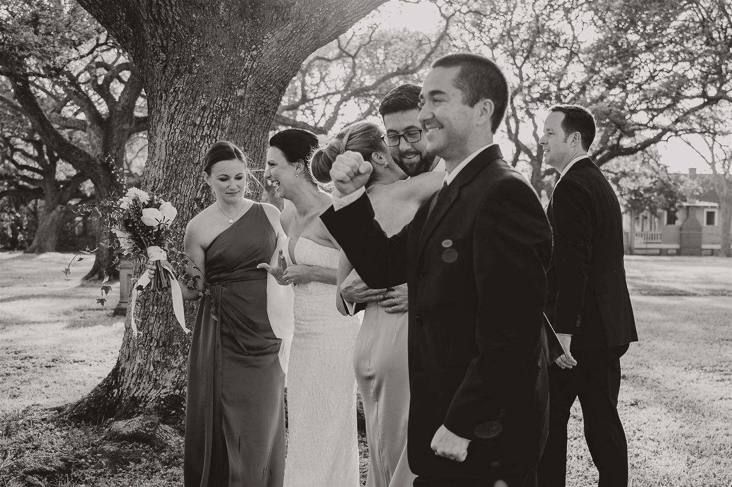 Wedding party interacts casually under trees during outdoor celebration.