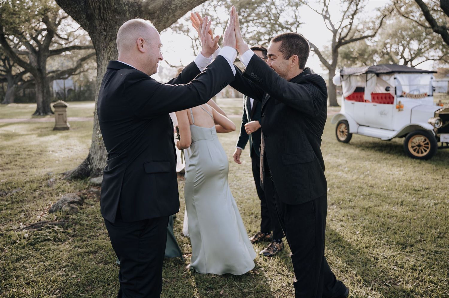 Groomsmen high-five during outdoor wedding celebration near vintage vehicles.