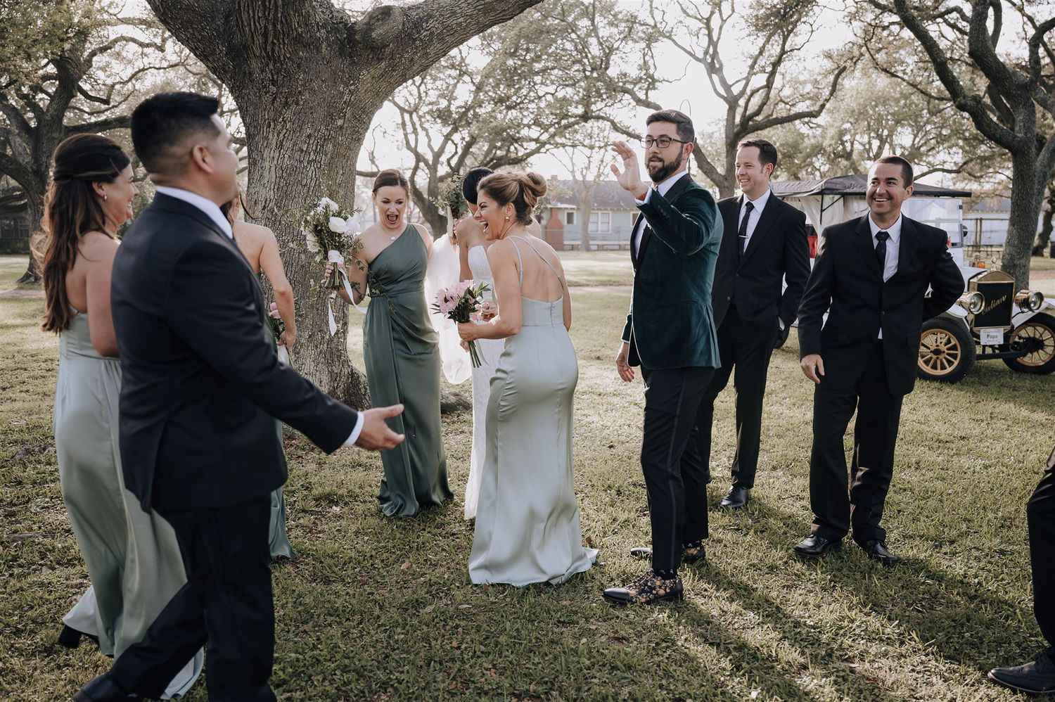 Wedding party gathers outdoors under large tree branches during a casual celebration.